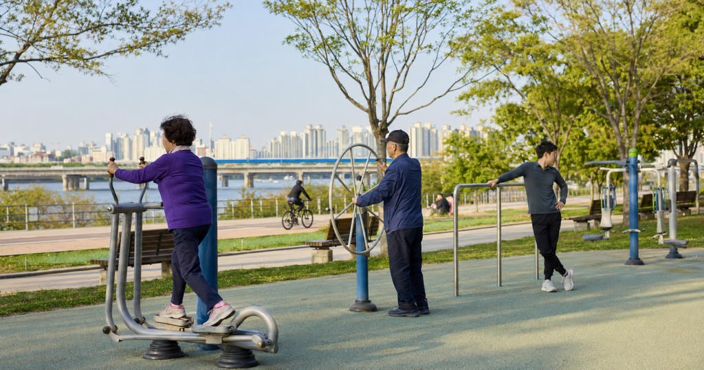 Local residents using free outdoor exercise equipment beside a riverside path in a Korean park.